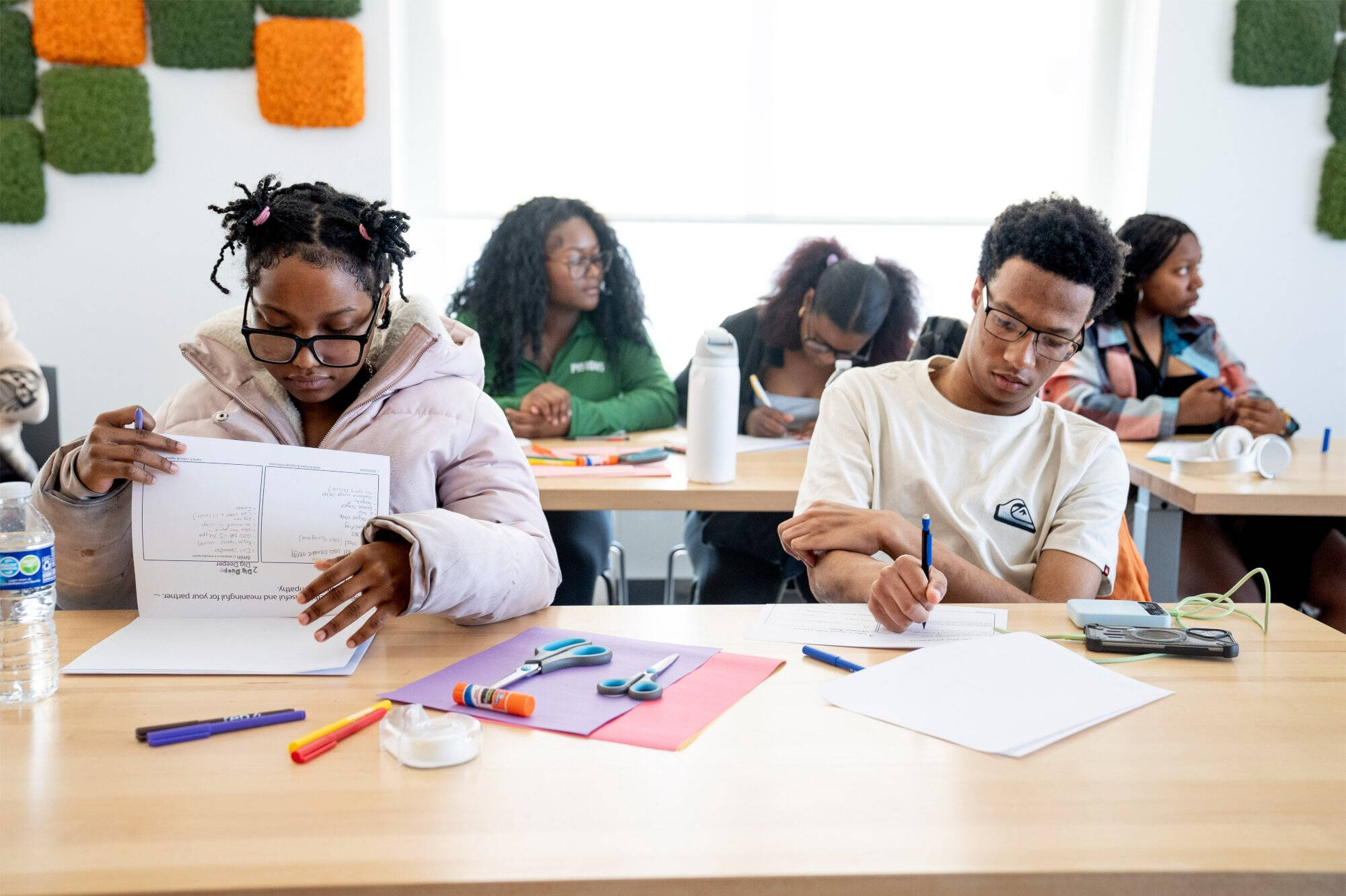 Students sit at tables working on a worksheet activity, using markers, scissors, and glue sticks to complete a paper folding exercise